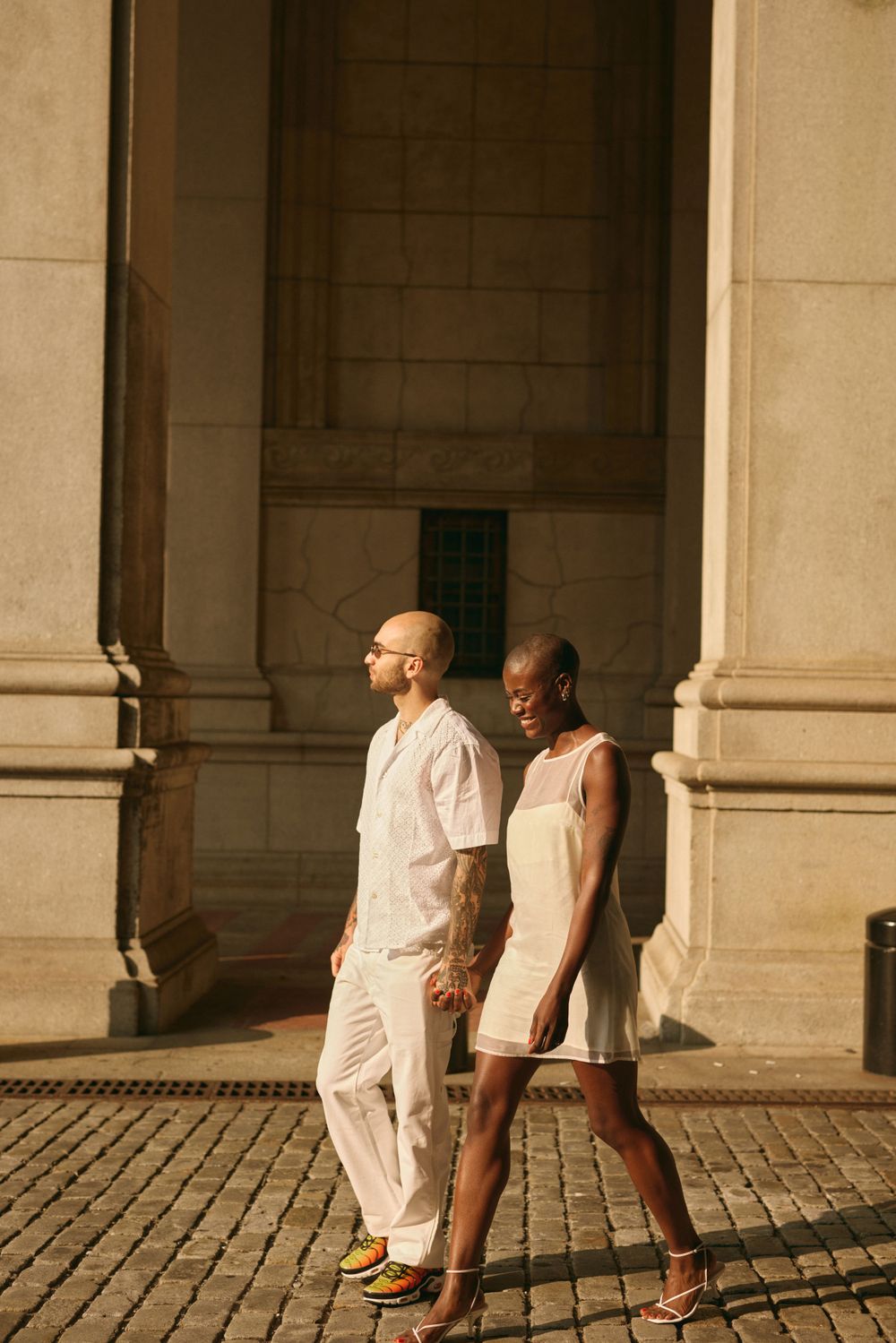Couple in light summer clothing walking on cobblestones near architectural columns.