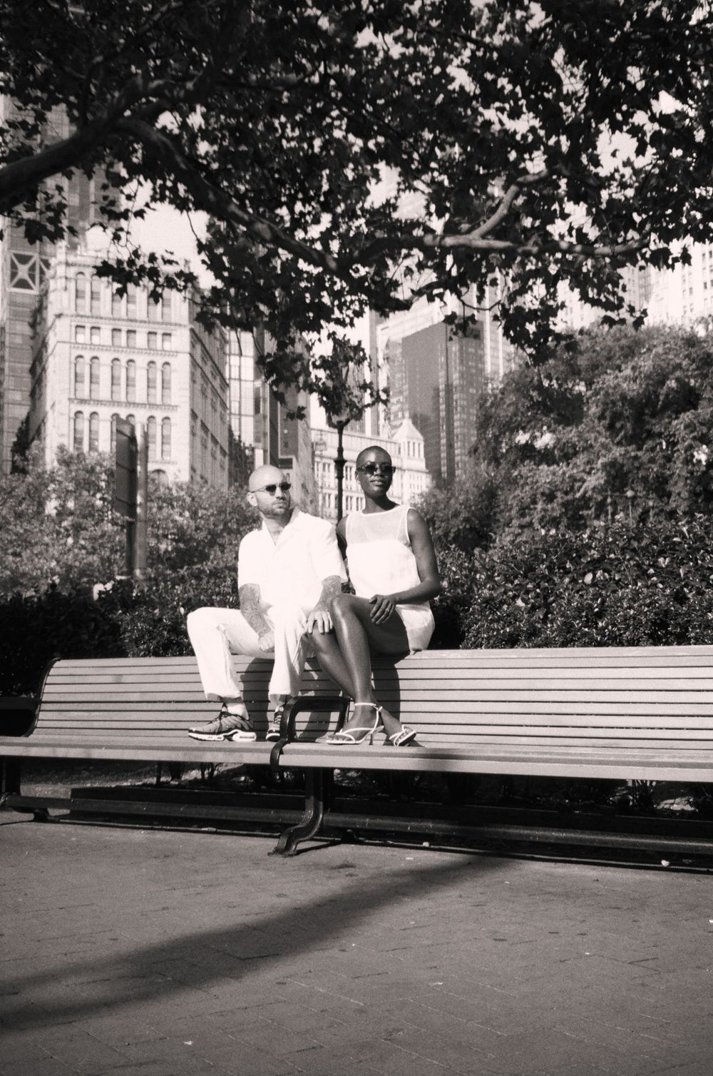 A couple relaxes on a park bench with urban buildings visible in the background.