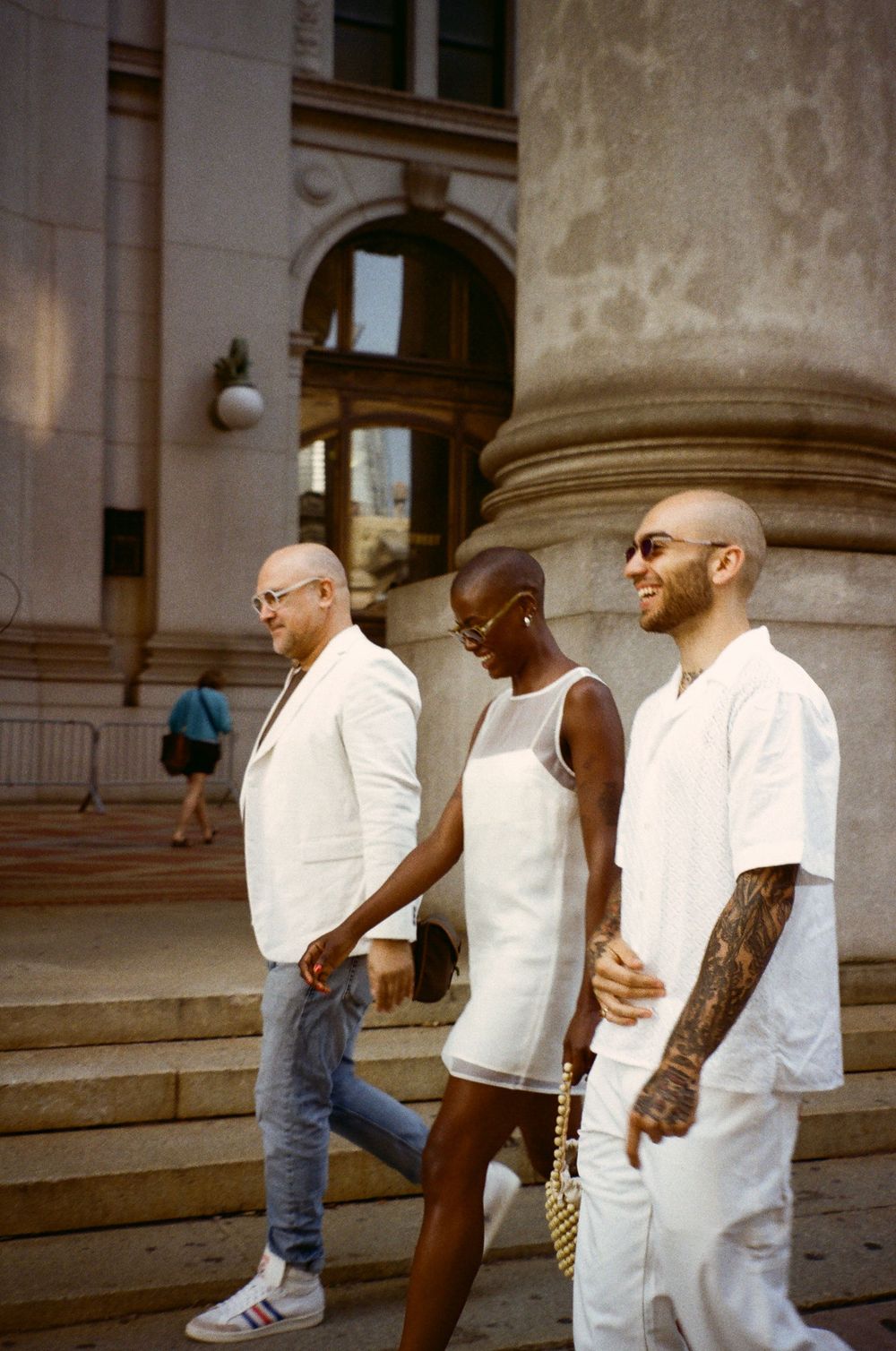 Three friends in coordinated white outfits walk down stone steps of a classical building on a sunny day.