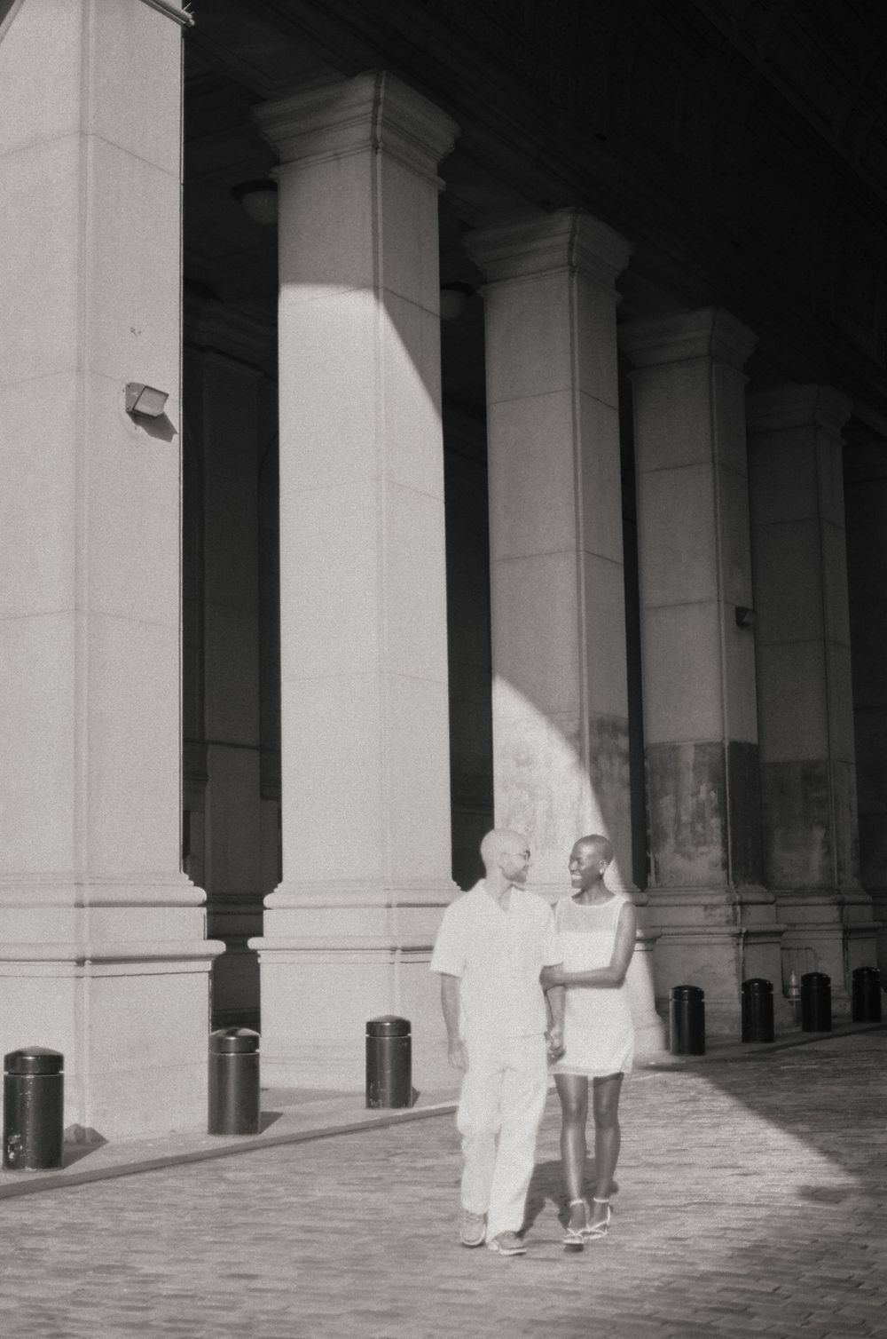 A couple in white outfits poses between classical white columns in black and white photography.