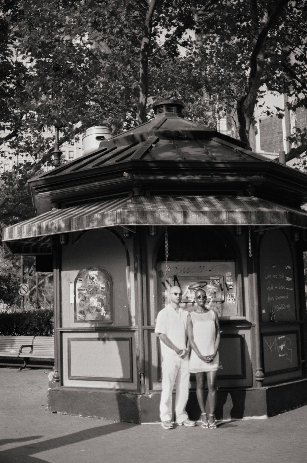 Two people pose together in front of a vintage hexagonal kiosk in a park setting.