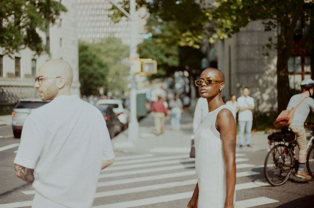People in white clothing walking on a city street with buildings and cyclists in the background.