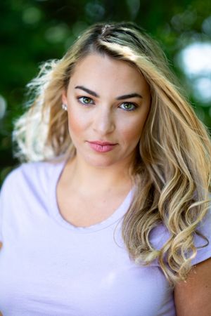 Portrait of a woman in lavender shirt with flowing blonde hair against green outdoor background.