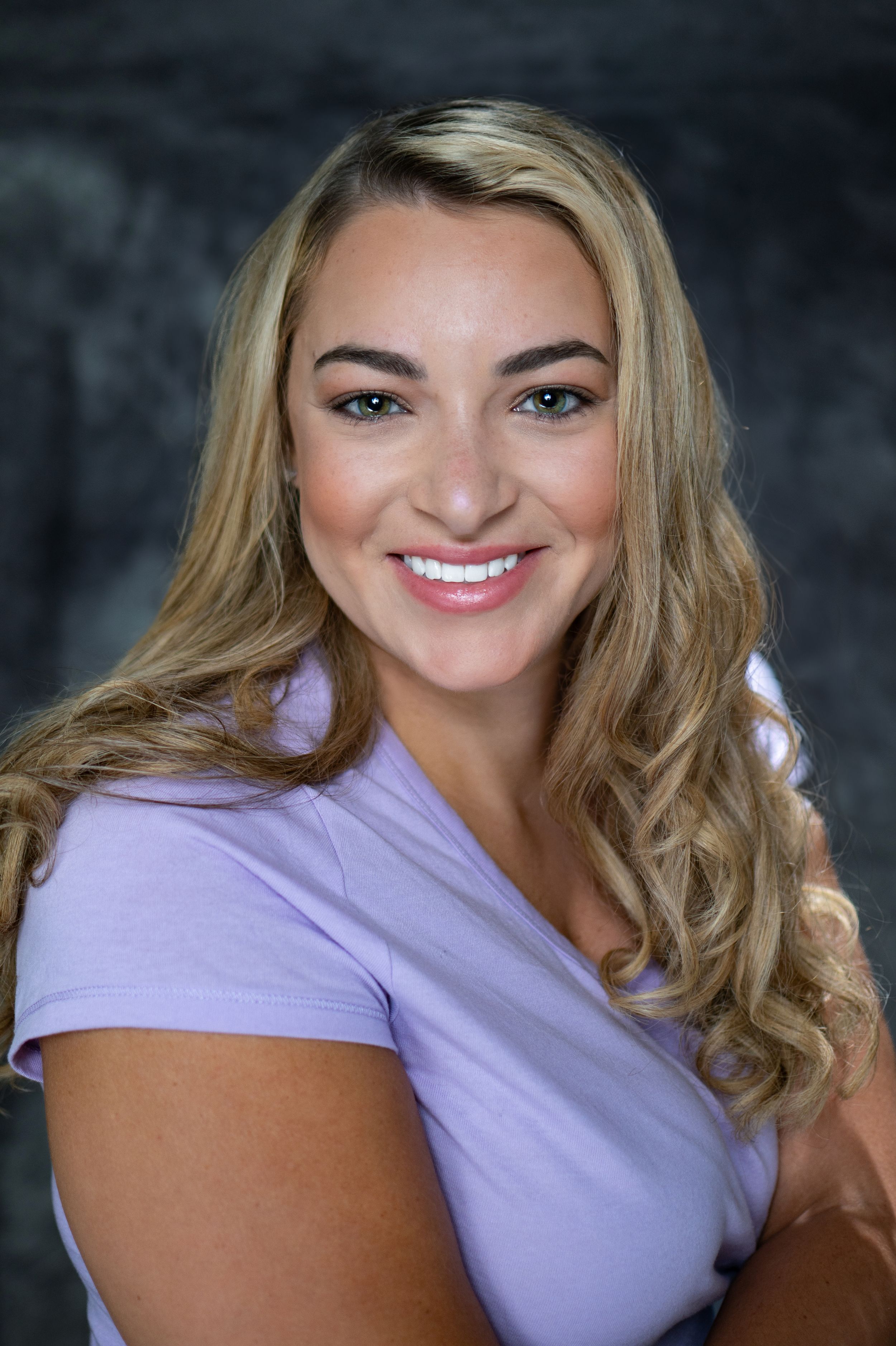 Professional headshot of a woman in a purple t-shirt smiling warmly against dark background.