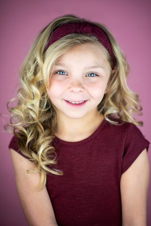 Portrait of child with curly blonde hair wearing burgundy top against pink background.