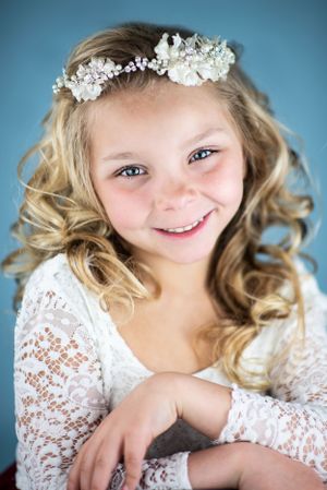 Child wearing lace dress and floral headpiece with curled hair against blue background.
