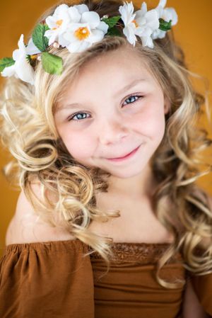 Young girl with curly blonde hair wearing a flower crown and brown dress against orange background.