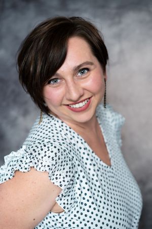 Professional headshot in polka dot blouse with short dark hair against grey background.
