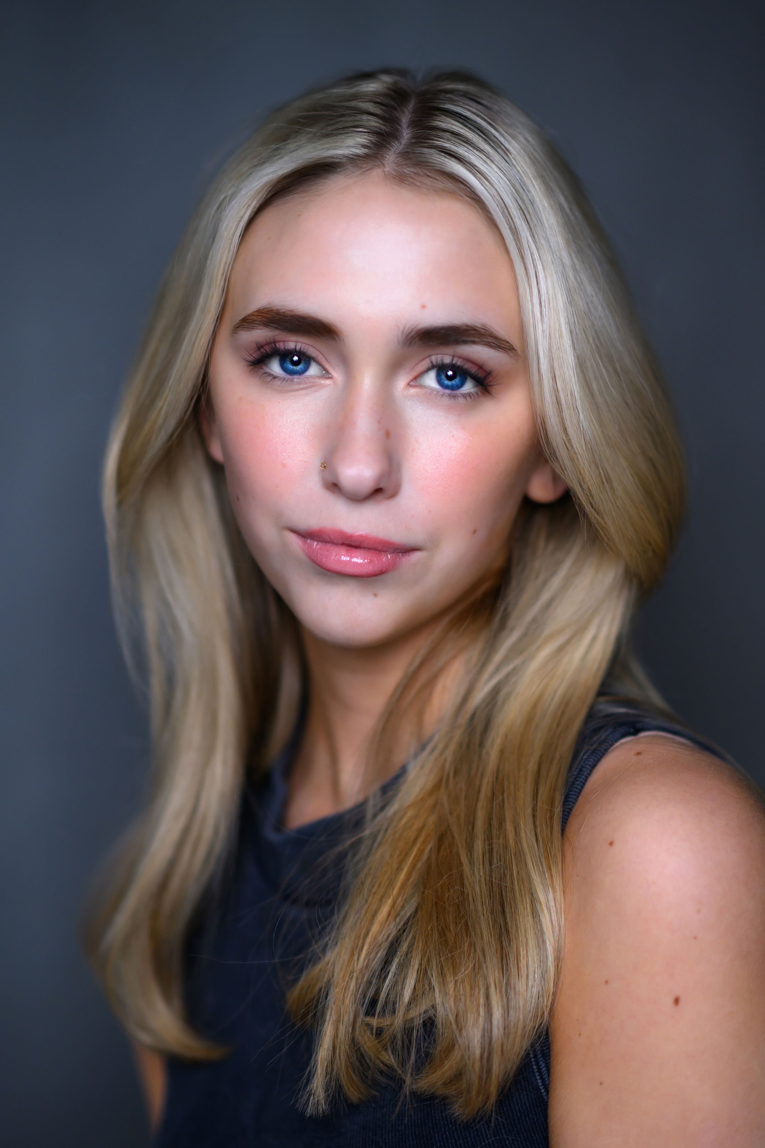 Professional headshot of a blonde model wearing a dark top against a gray background.