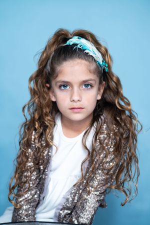 Stylish portrait with long curly hair, turquoise headband and sparkly jacket against blue background.