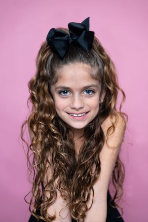 Portrait with long curly brown hair and black bow against pink background.