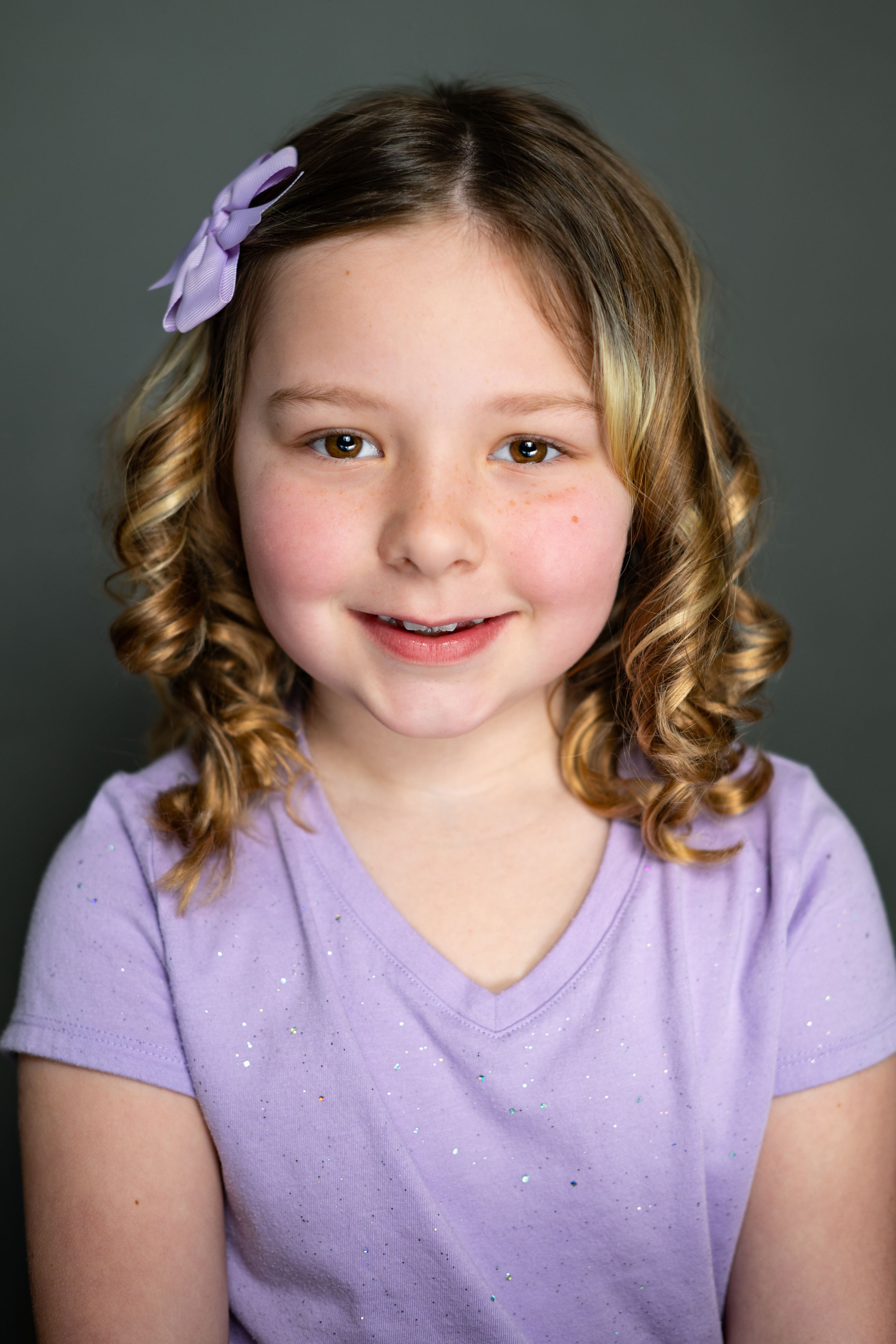 Portrait of a young girl in a purple shirt with curly blonde hair and a matching purple hair bow.