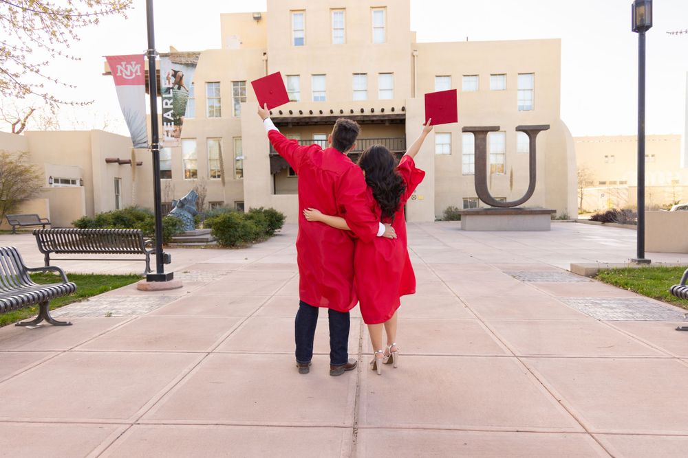 Two graduates in red caps and gowns celebrating with raised arms on campus.