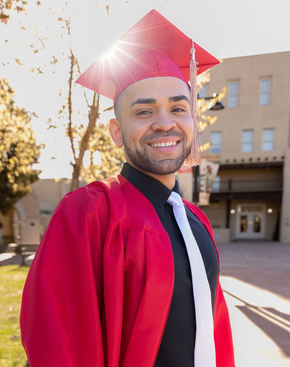 Graduate in red cap and gown smiles brightly against sunny campus building.