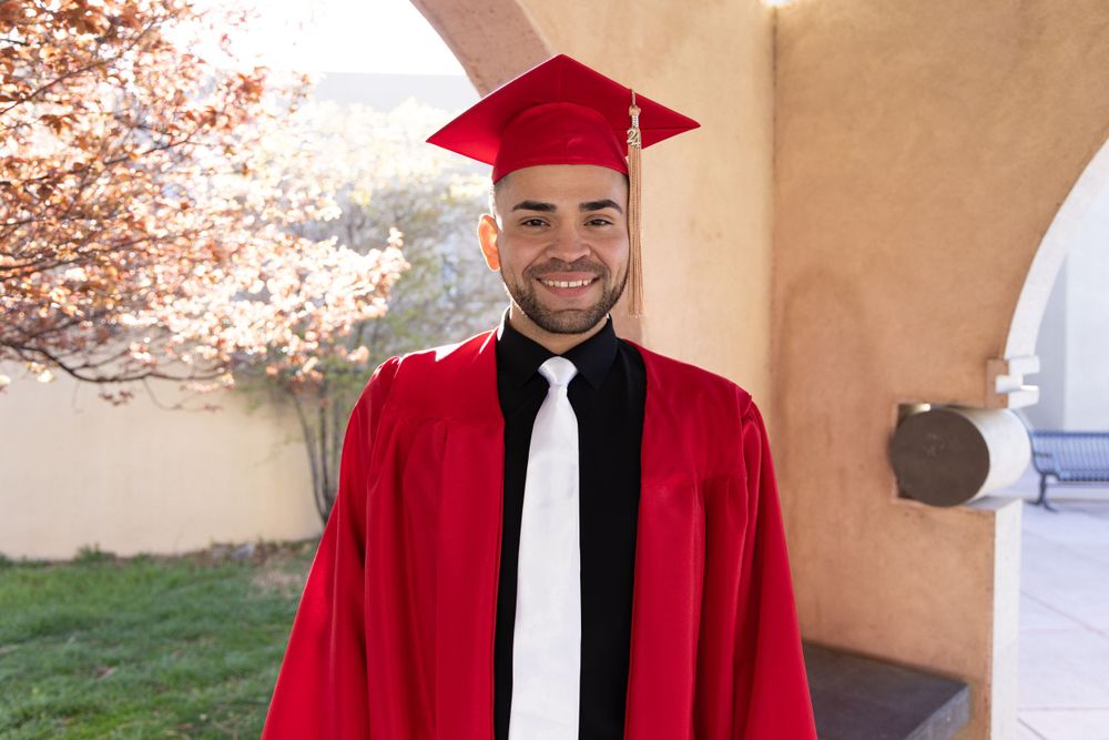 Graduate in red cap and gown with black tie smiles against adobe wall.