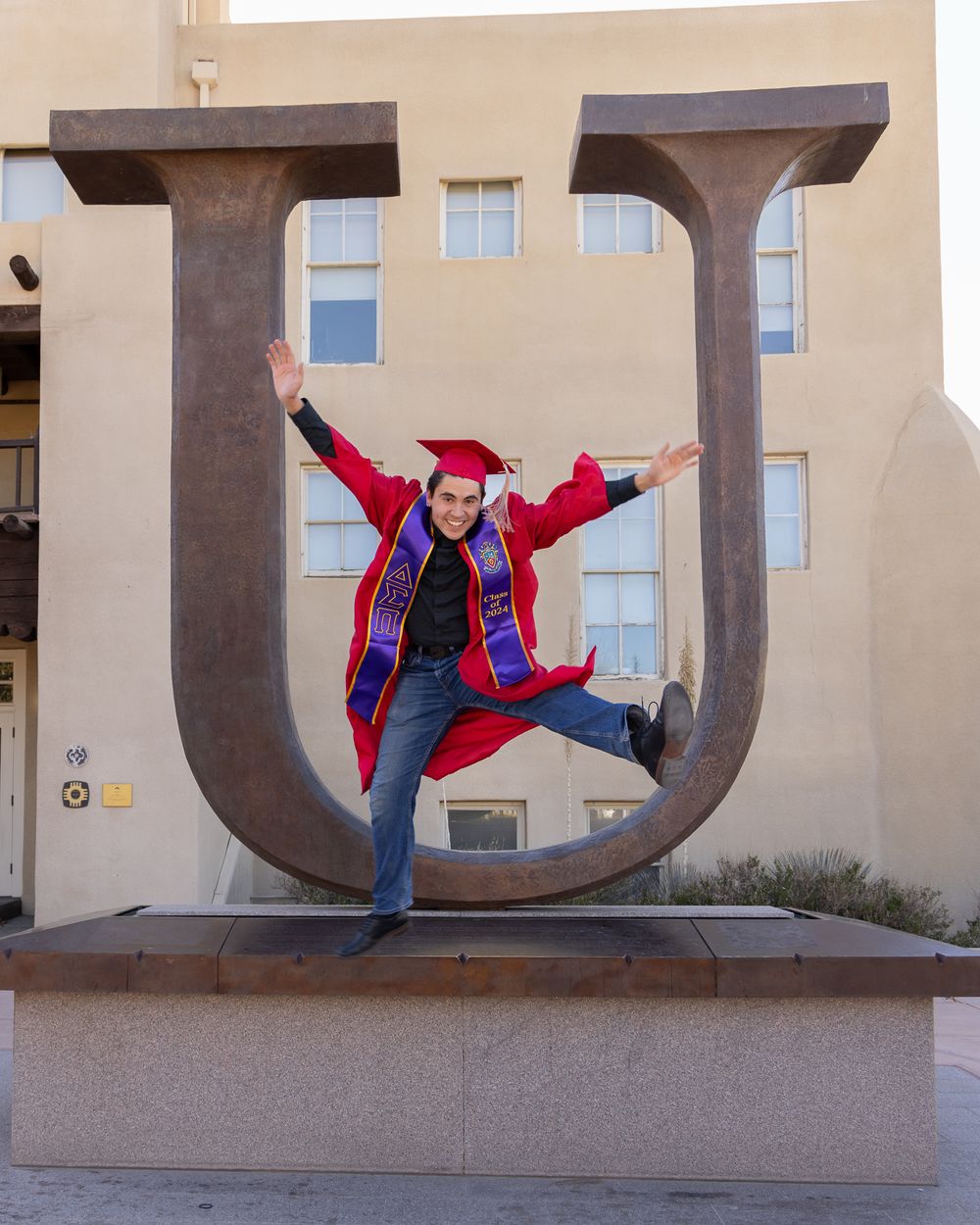 Graduate in red cap and gown jumps playfully in front of large U-shaped sculpture.