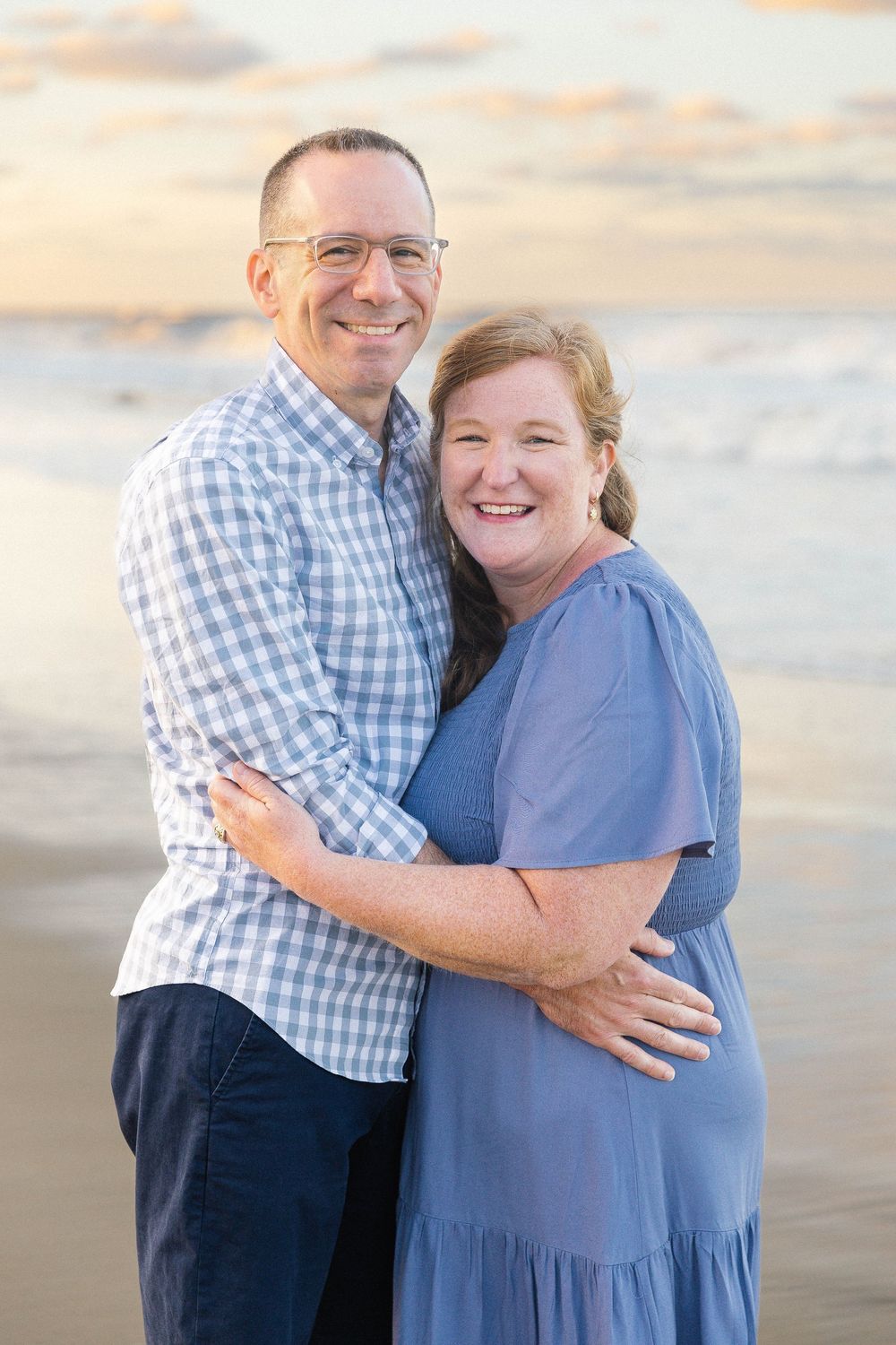 Couple embracing on beach at sunset wearing blue and plaid clothing.