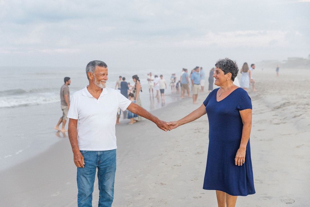 Couple walking and holding hands on beach with others in background.