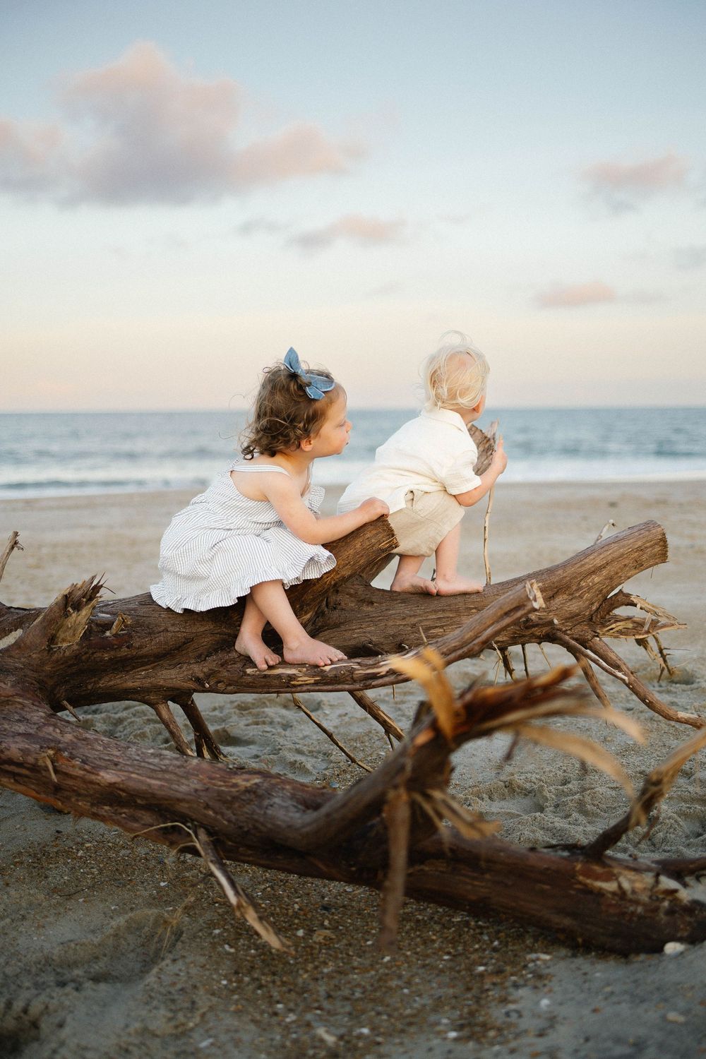 Two young children sit on driftwood at a beach during sunset, wearing white outfits, gazing at the ocean.