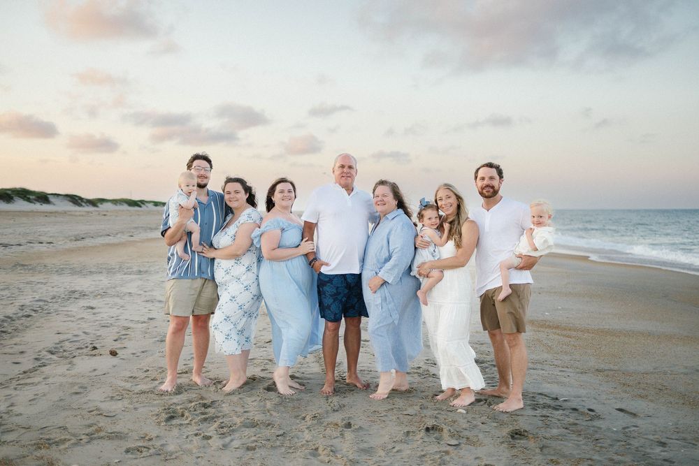 Family of eight poses together on a sandy beach at sunset wearing coordinated blue and white outfits.