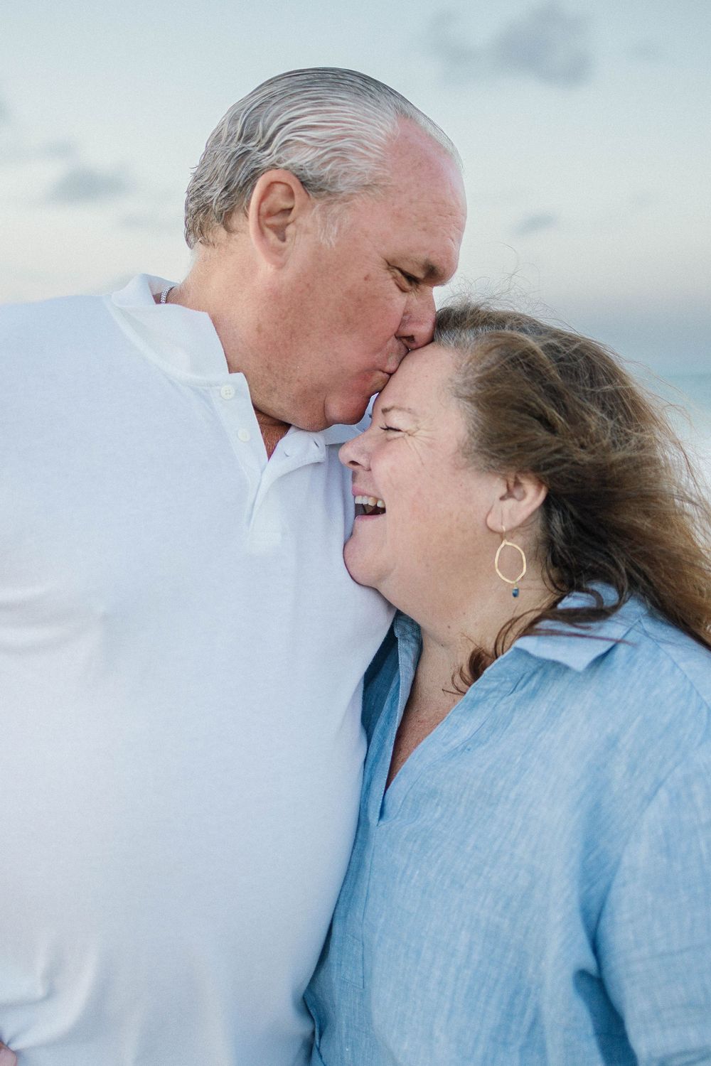 An older couple shares a tender moment on the beach, with the man kissing the woman's forehead as she laughs joyfully.