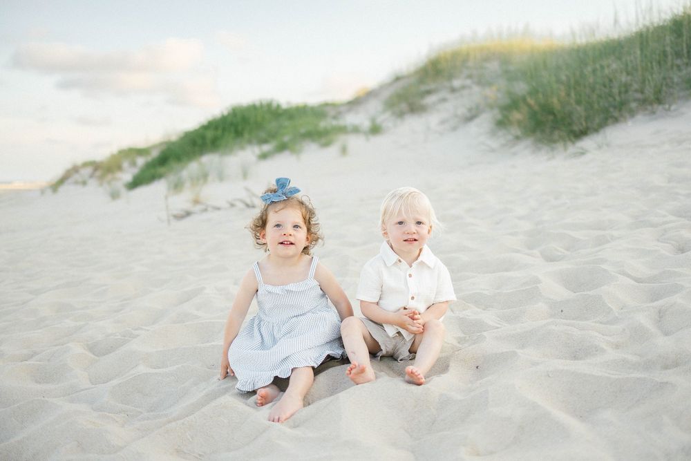 Two young children sitting on sandy beach dunes, girl in blue dress and boy in white shirt, smiling outdoors.