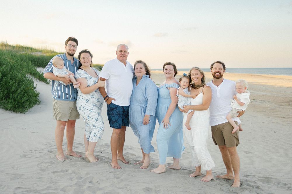 Multi-generational family posing together on a sandy beach at sunset, smiling in coordinated blue and white outfits.