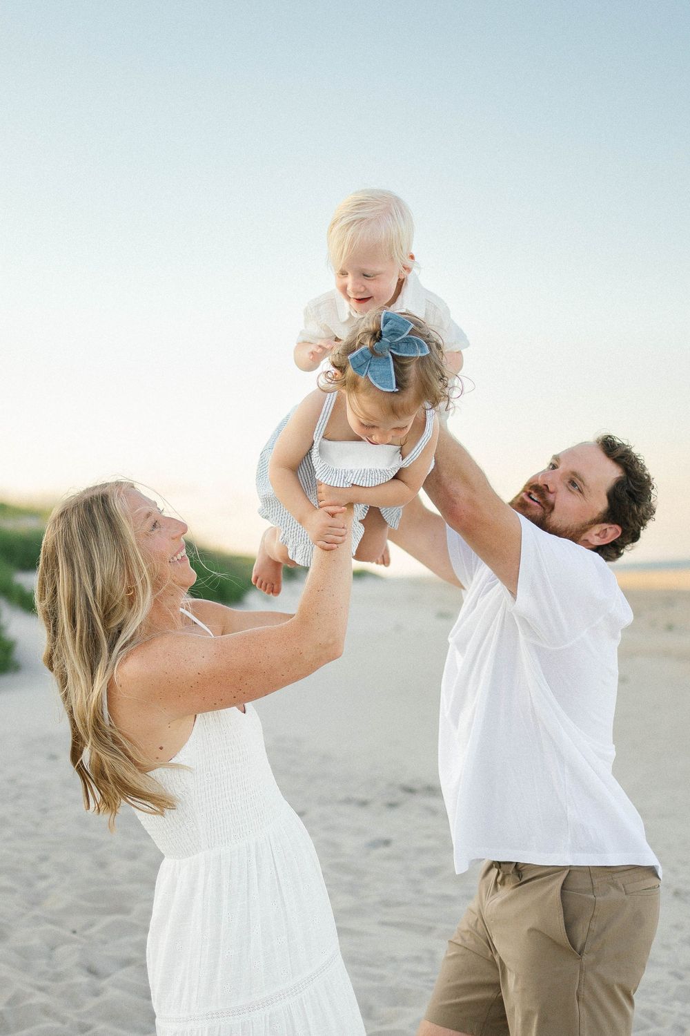 A happy family plays on a sandy beach at sunset, with parents lifting their laughing toddler in the air.