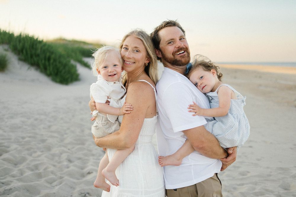 Happy family of four posing on a sandy beach at sunset, parents holding two young children in white summer outfits.