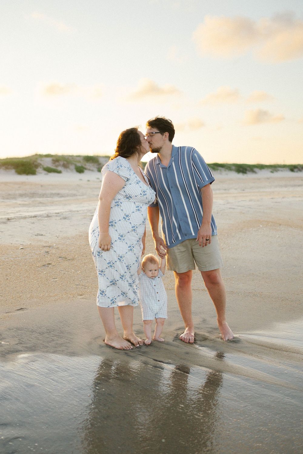 A couple kisses on a sandy beach at sunset while their toddler stands between them near the water.