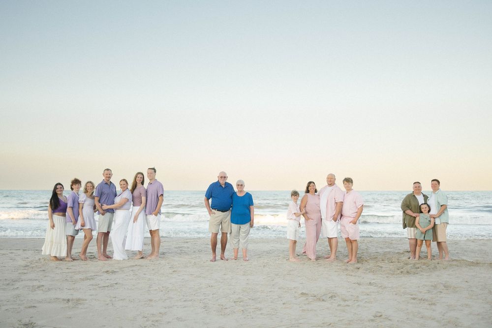Large family group portrait on a sandy beach at sunset, dressed in coordinating pastel outfits with ocean waves in the background.