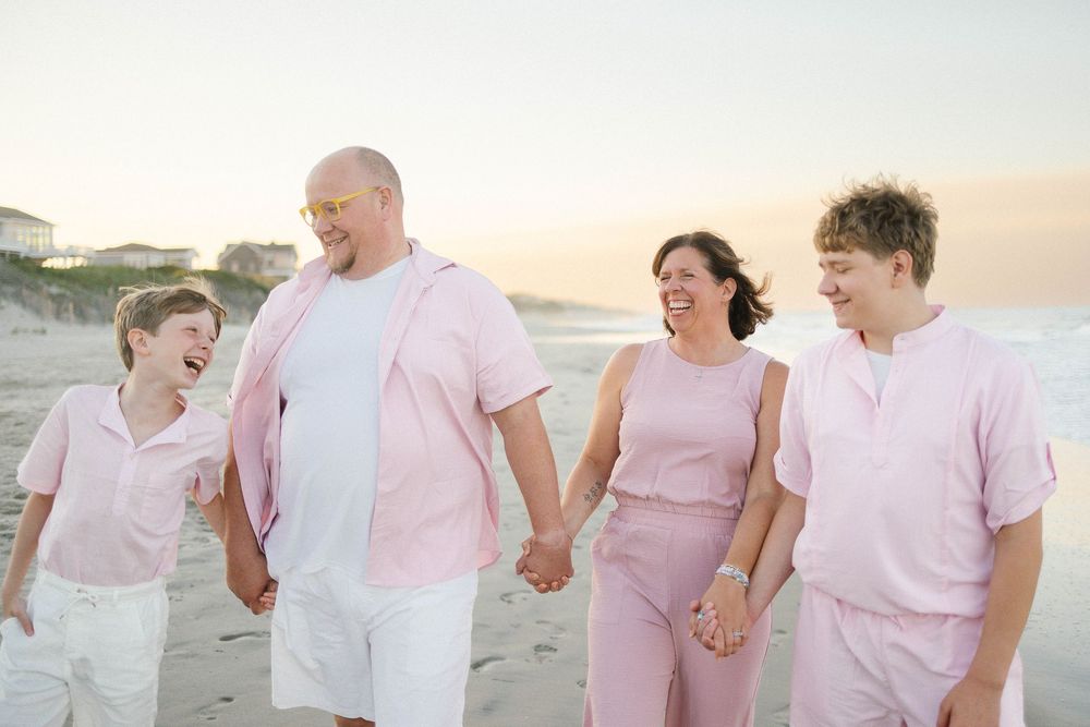 Family of four in matching pink outfits holding hands and laughing on a beach at sunset.