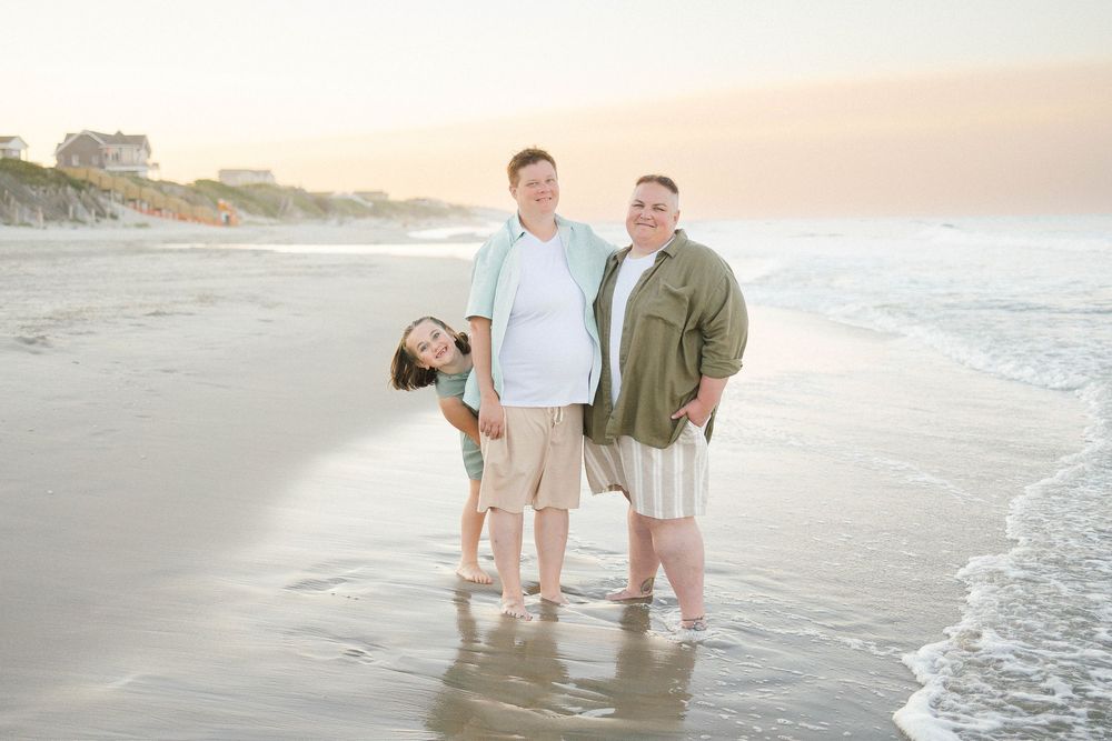 Family of three posing on a sandy beach at sunset, waves gently washing ashore behind them.
