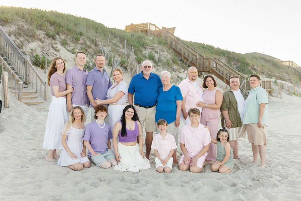 Large family group portrait on sandy beach at sunset, wearing coordinated pastel outfits, with dunes and wooden stairs behind them.