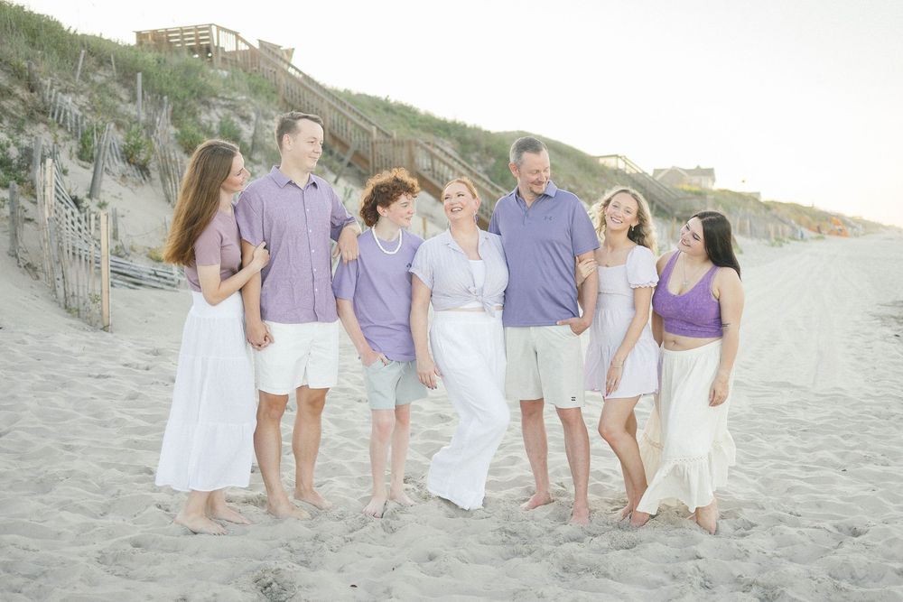Family of seven in coordinating purple and white outfits laughing together on a sandy beach at sunset.