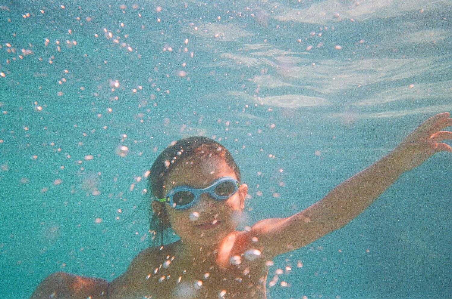 Underwater photo showing someone swimming with blue goggles and bubbles in turquoise water.