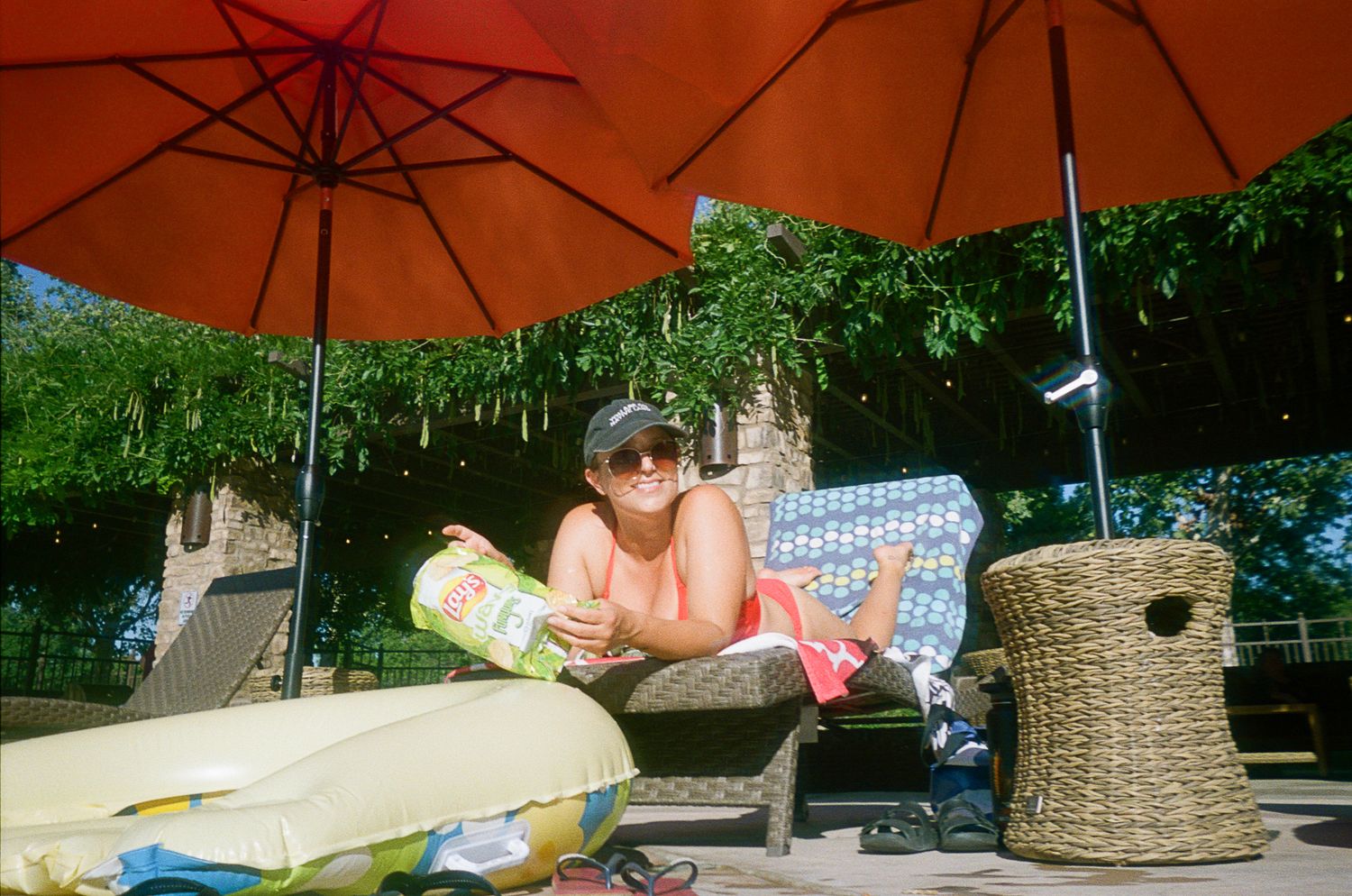 Person relaxes on an outdoor patio under an orange umbrella with cushions and a woven basket.