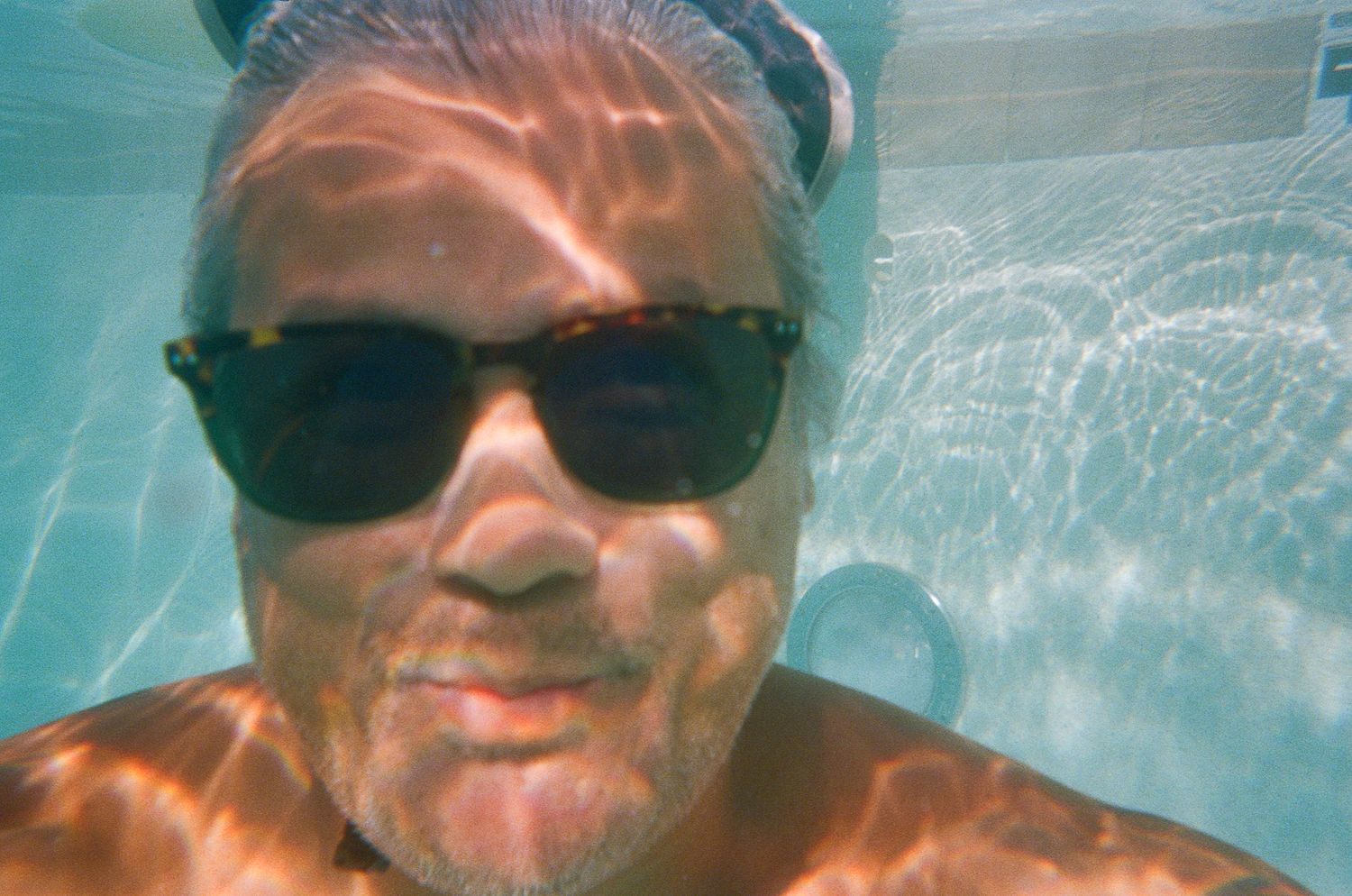 Underwater selfie showing a swimmer wearing sunglasses in a pool with sunlit ripples.
