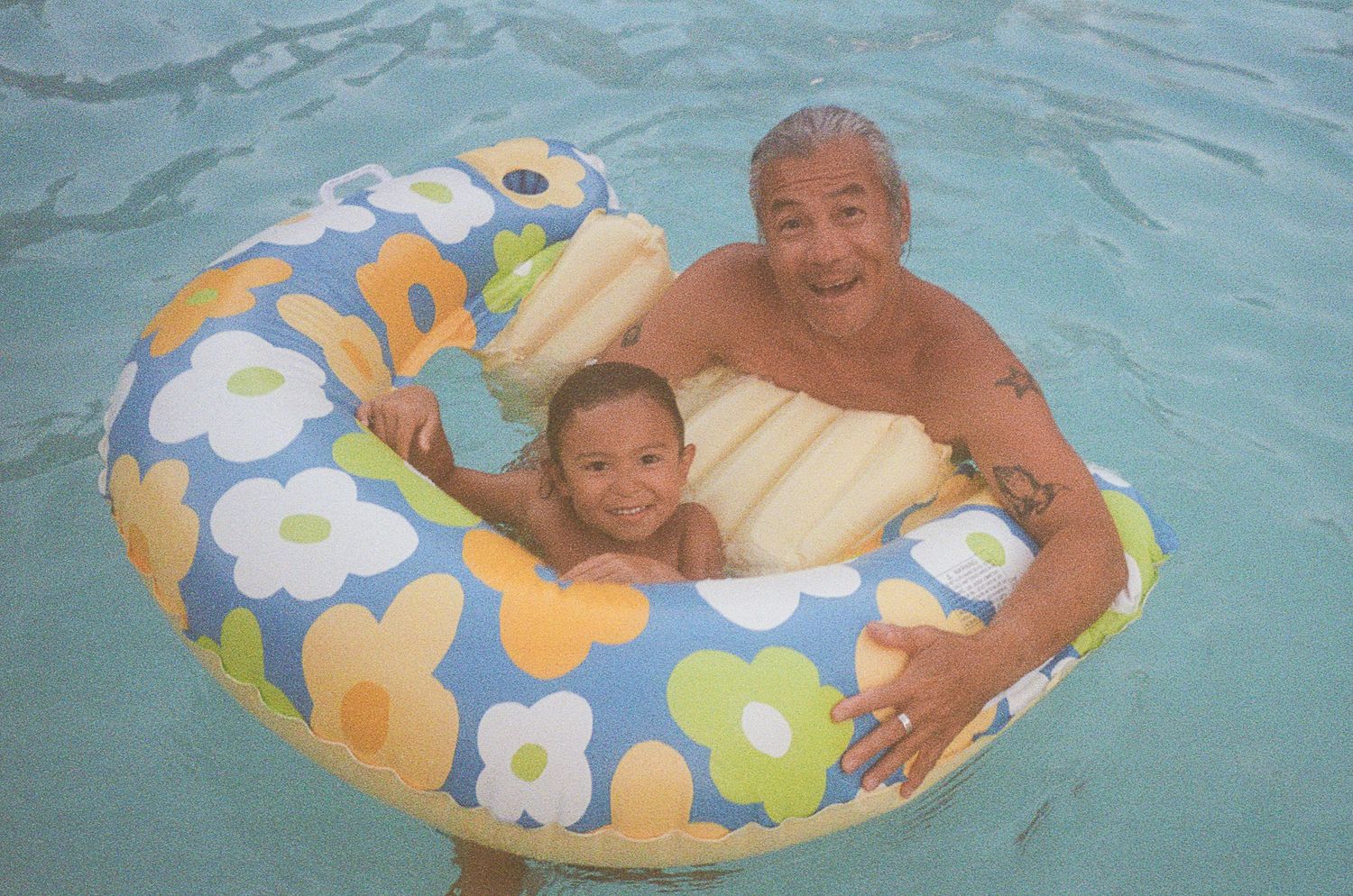 Two swimmers share a colorful floral pool float in turquoise water.