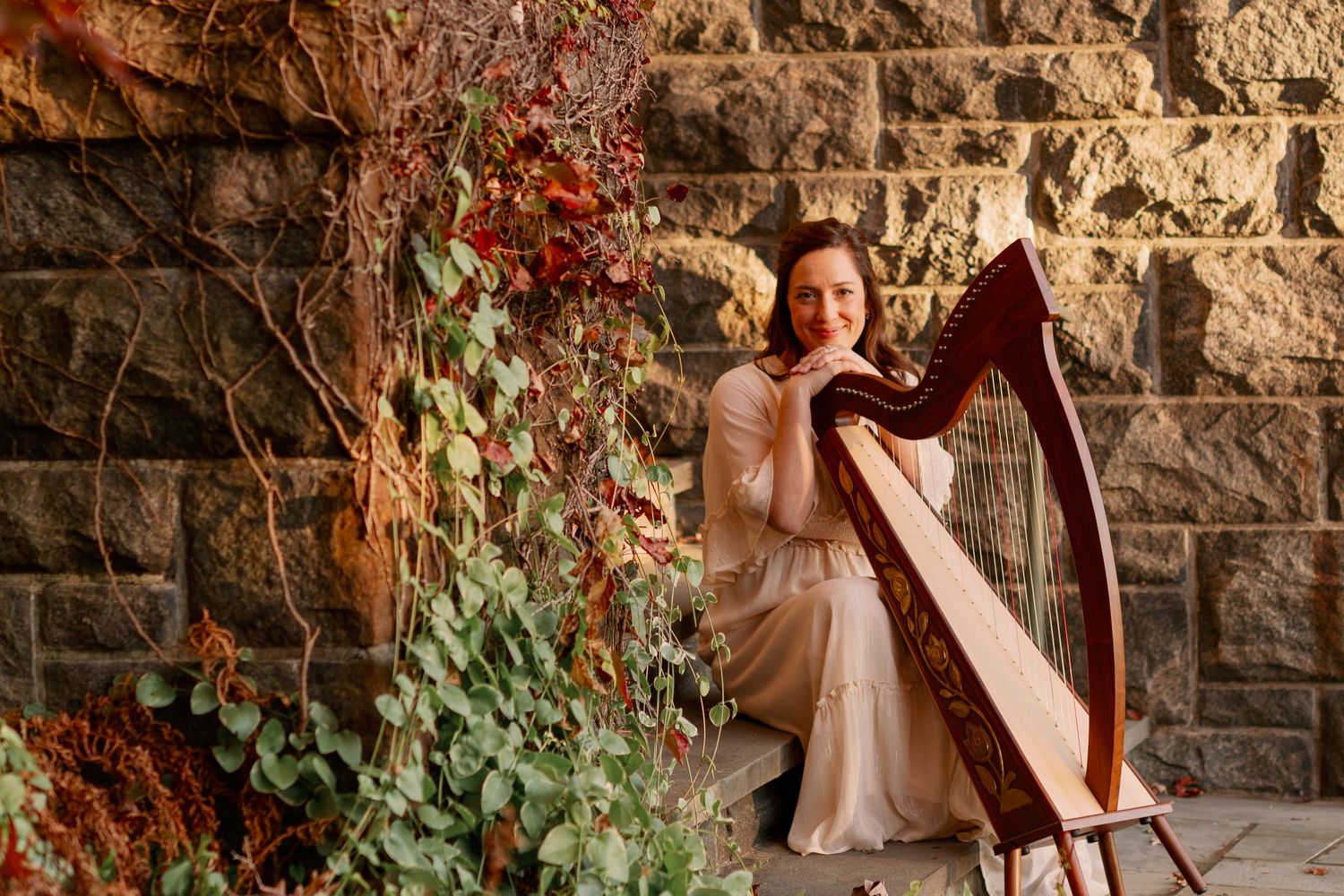 A harpist in an elegant dress performs beside a stone wall with climbing red ivy.