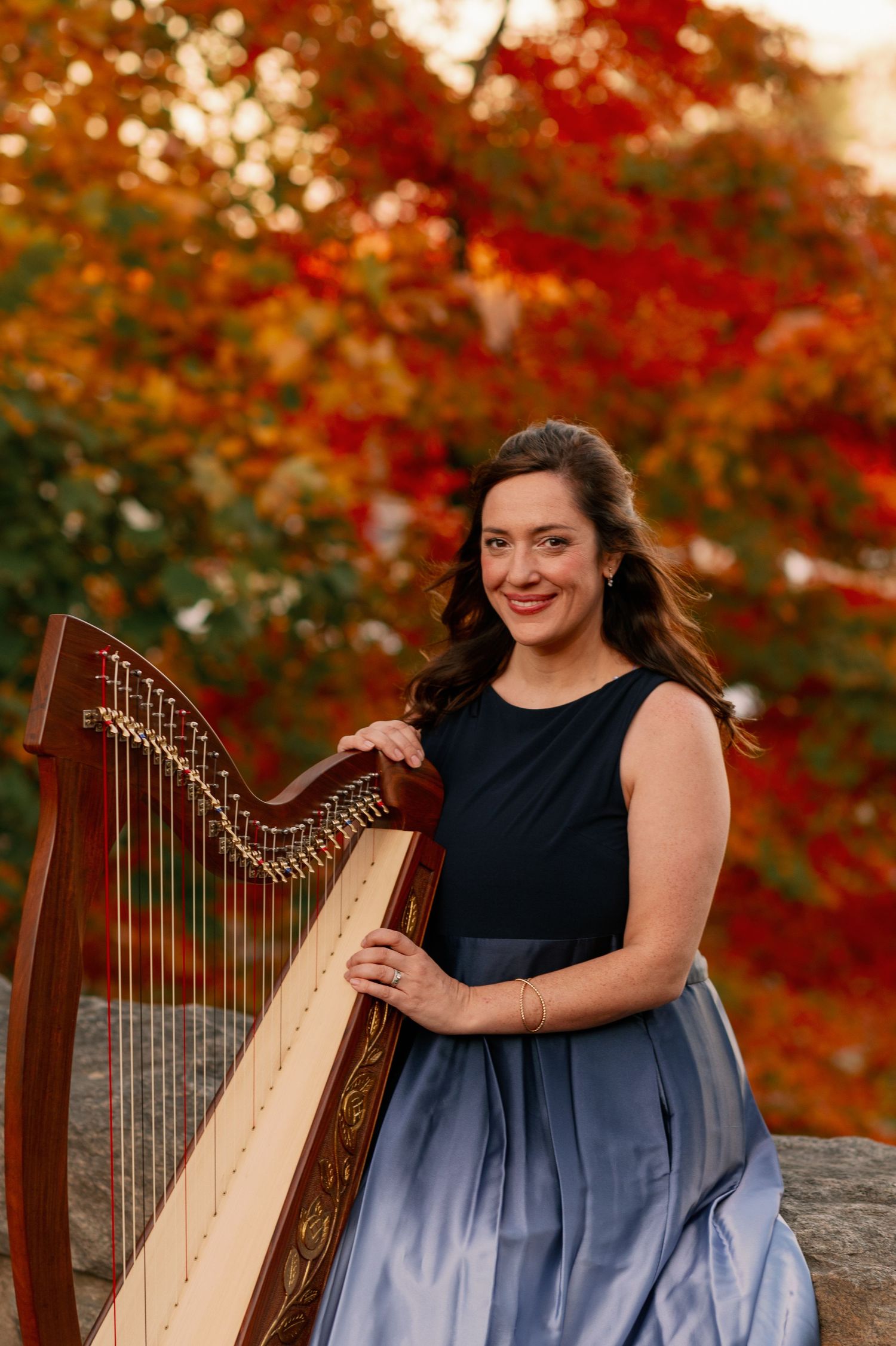 A harpist in a navy blue gown performs outdoors with autumn colors in the background.