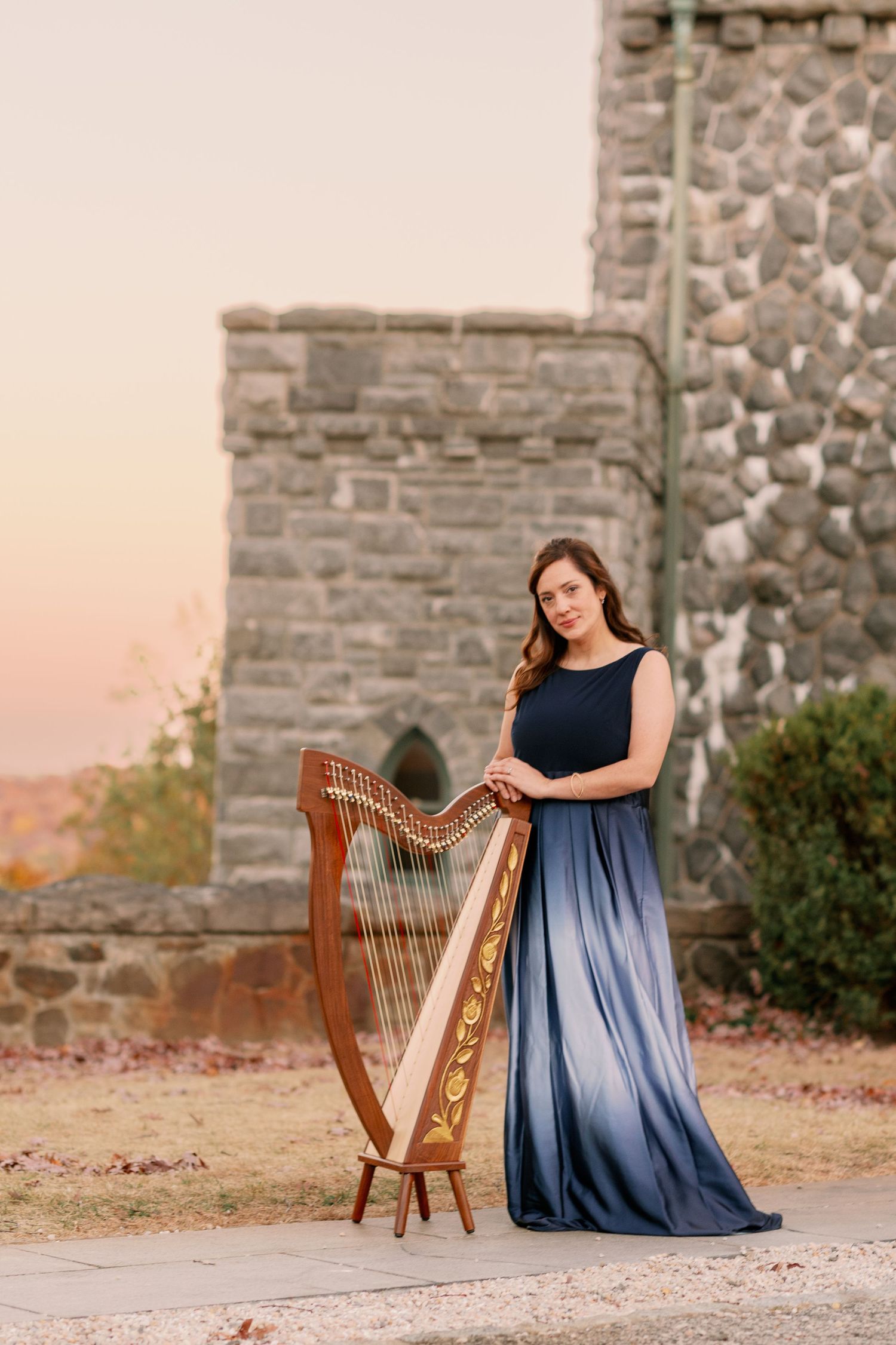 A harpist in a flowing blue dress performs near stone castle walls at sunset.