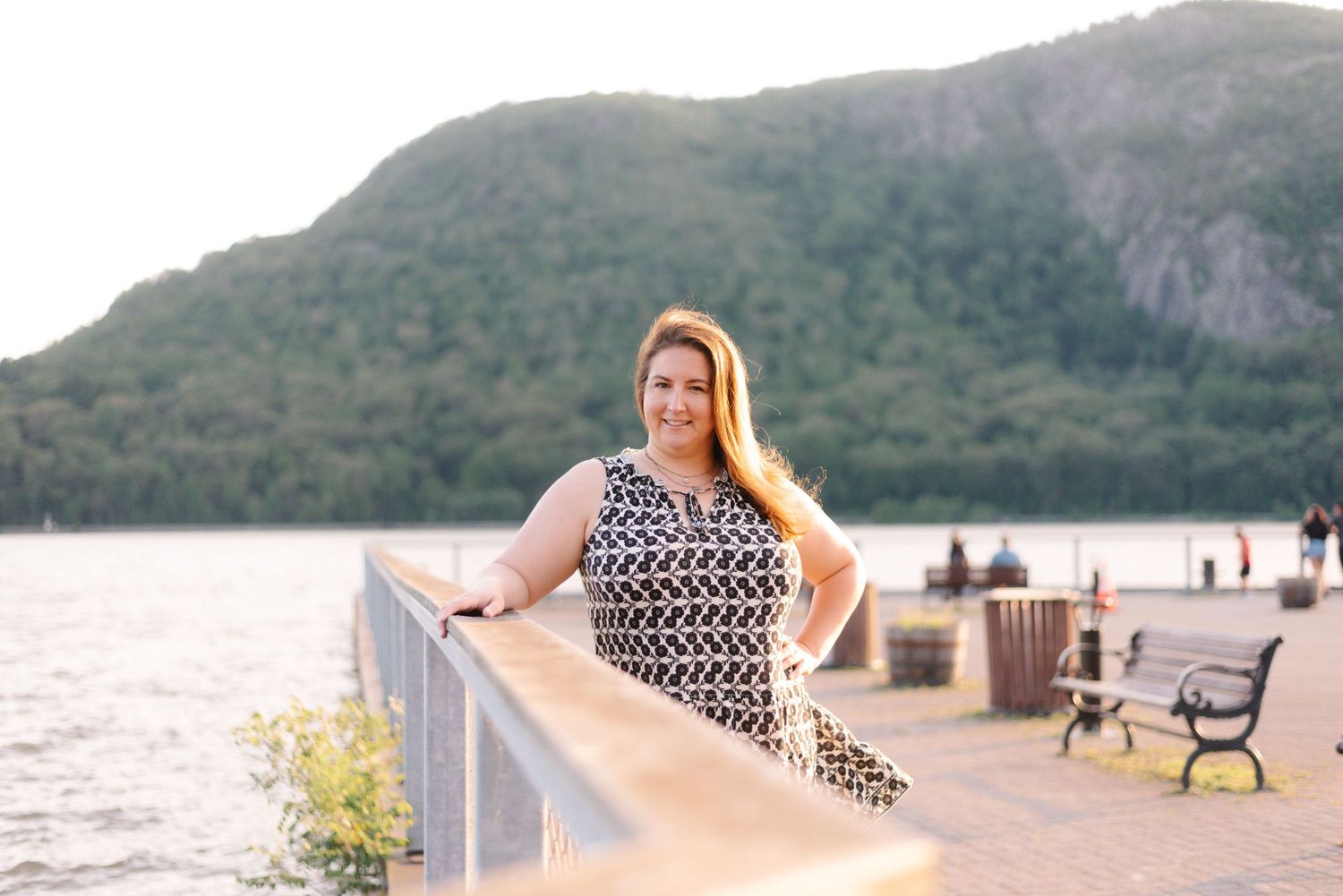 Woman in patterned dress poses on waterfront boardwalk with mountain landscape at sunset.