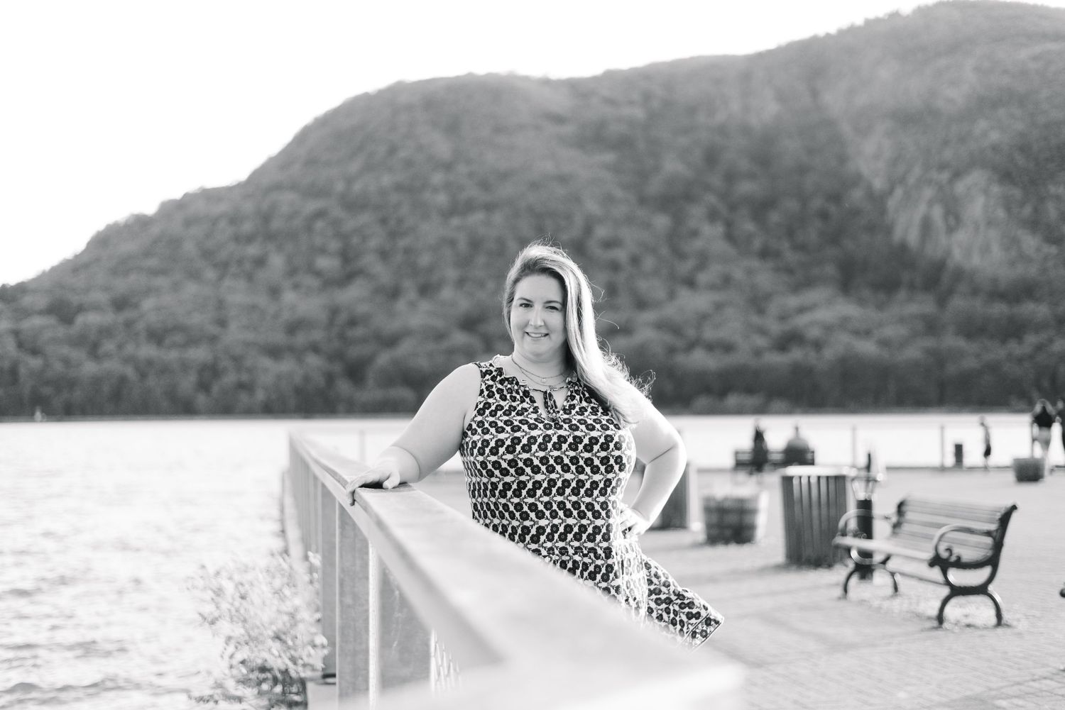 Black and white portrait of woman in patterned dress on waterfront boardwalk with mountain backdrop.