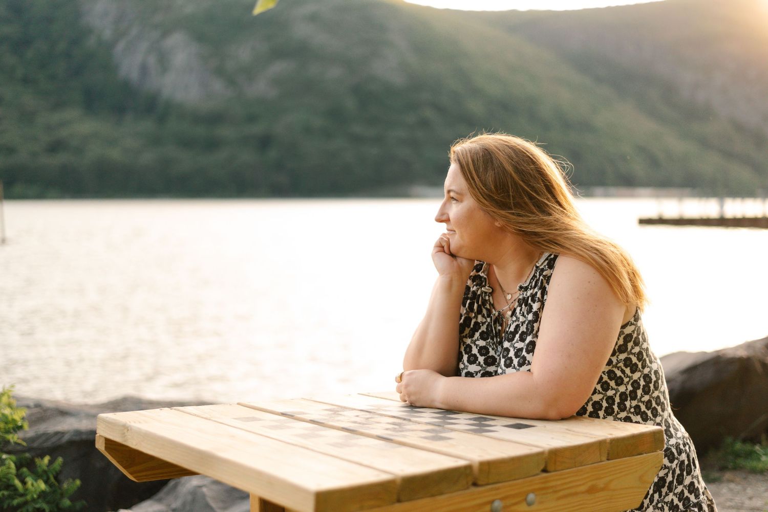 Woman in patterned dress relaxes at wooden table by lakeside during golden hour.
