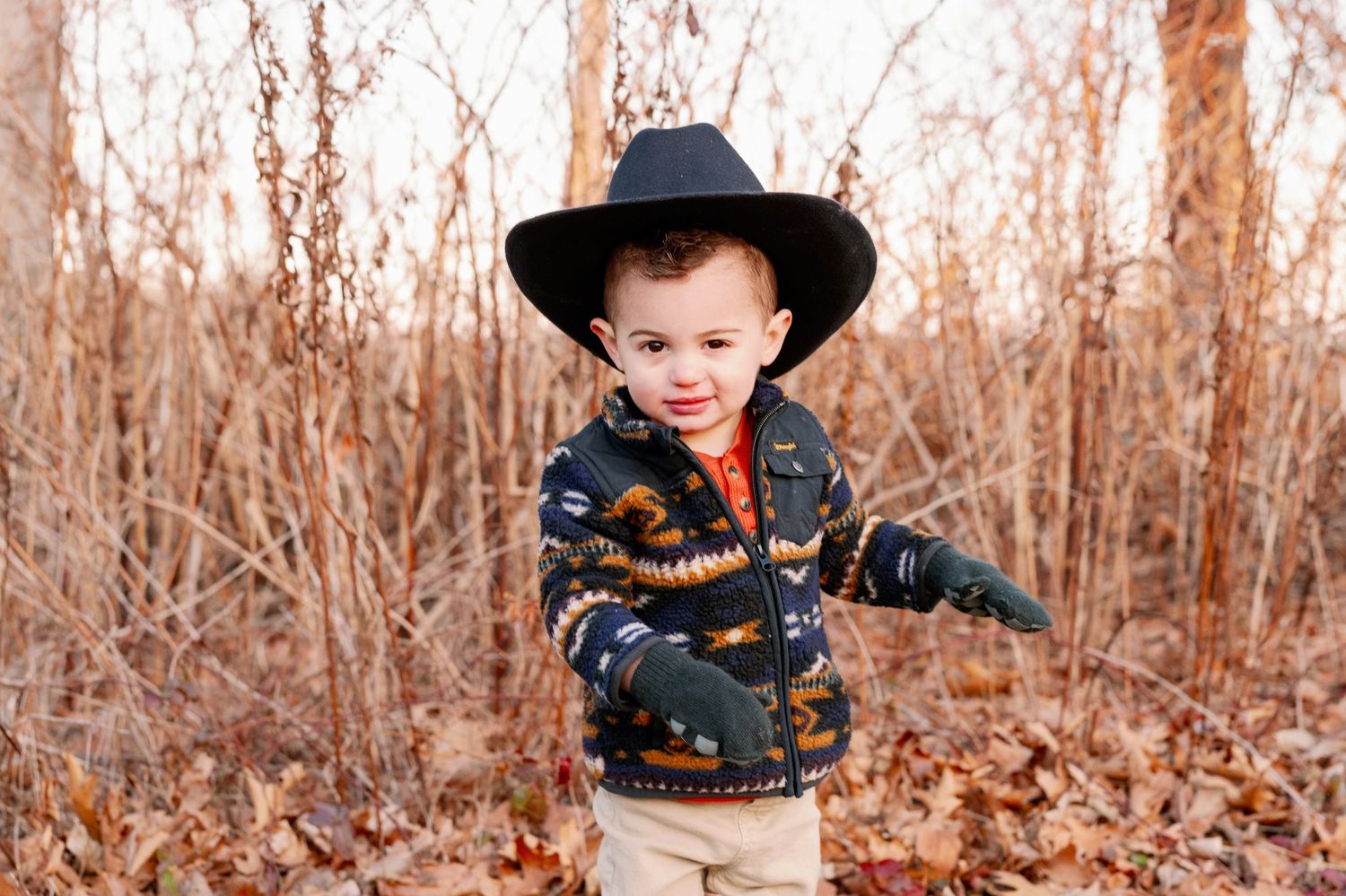 A small child wearing a cowboy hat and western-style jacket stands in tall autumn grass.