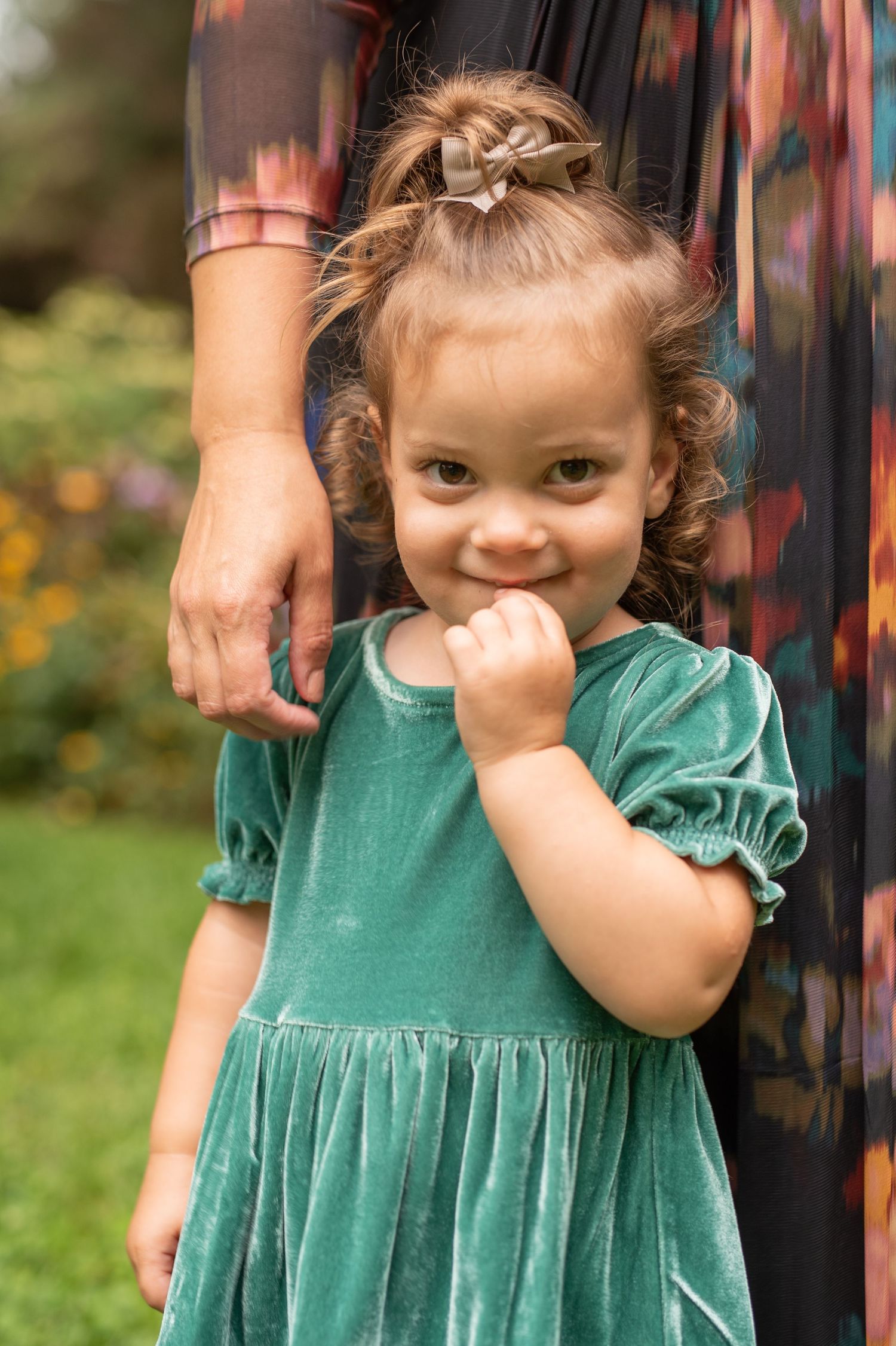 A toddler in a teal green dress smiles shyly while standing next to an adult.