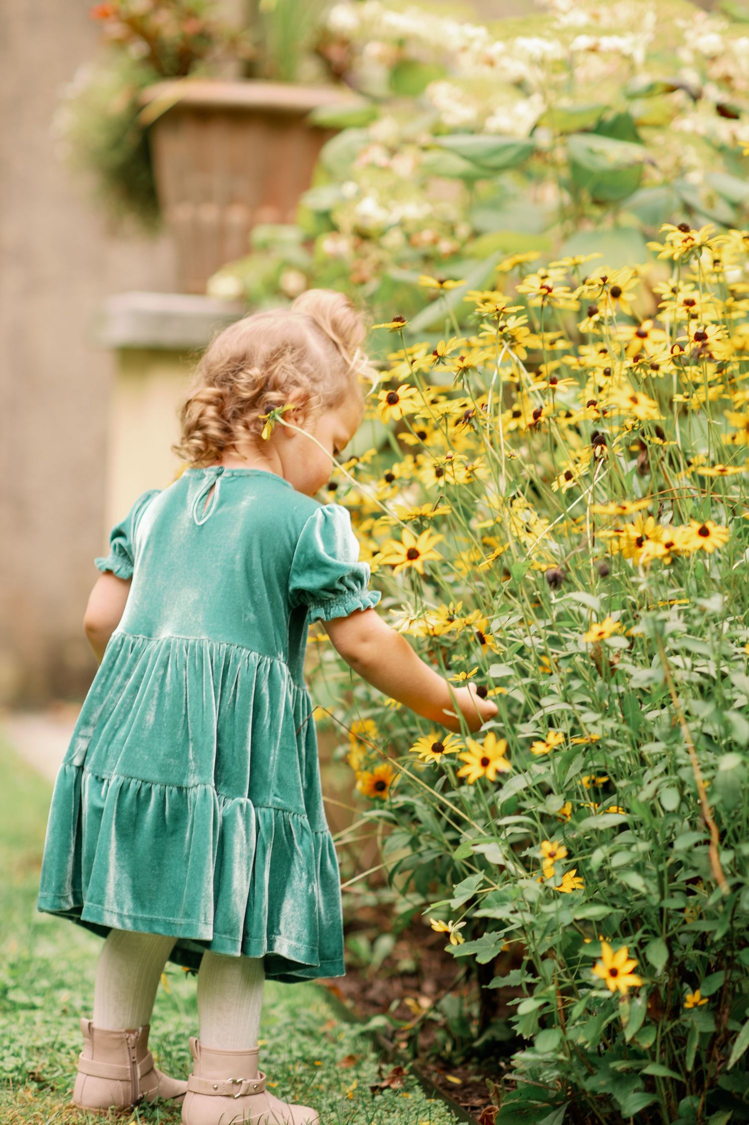 A child in a teal dress explores a garden filled with blooming yellow flowers on a sunny day.
