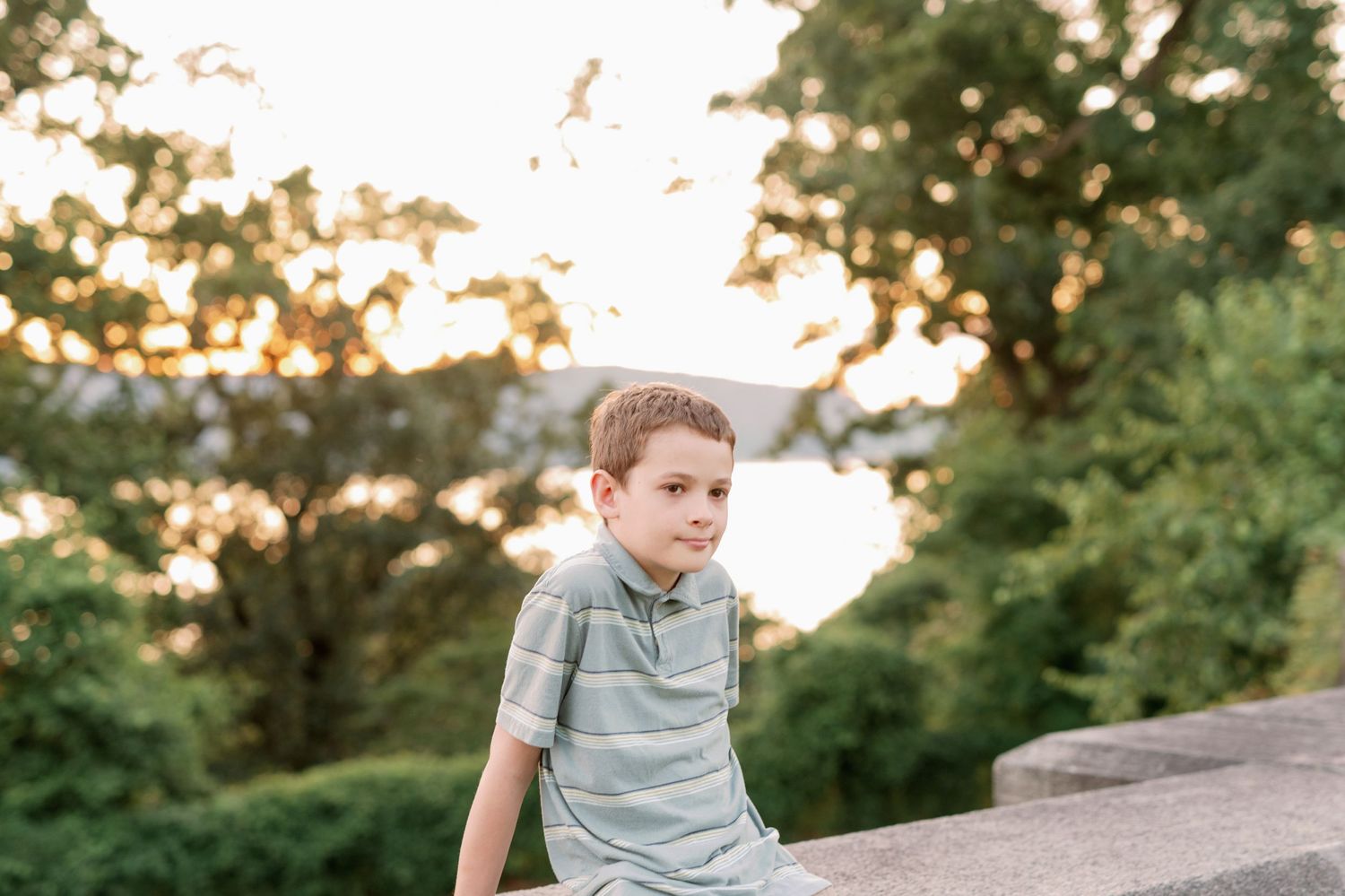 Boy in striped shirt sits on stone wall during golden hour photoshoot.
