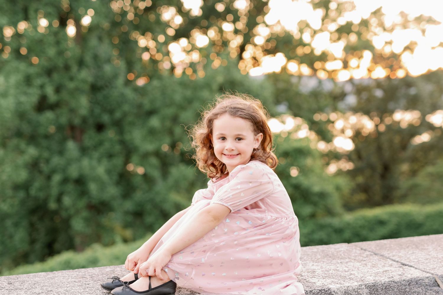 Girl in pink dress sits on stone wall during sunset outdoor portrait session.